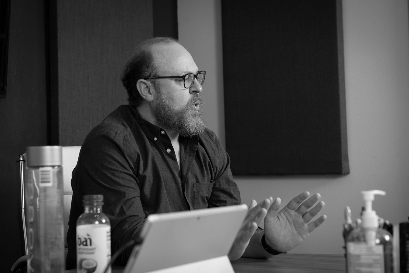 Black and white photo of Max Kozlovsky seated at a conference table, speaking with his hands open, a tablet and water bottles in front of him.