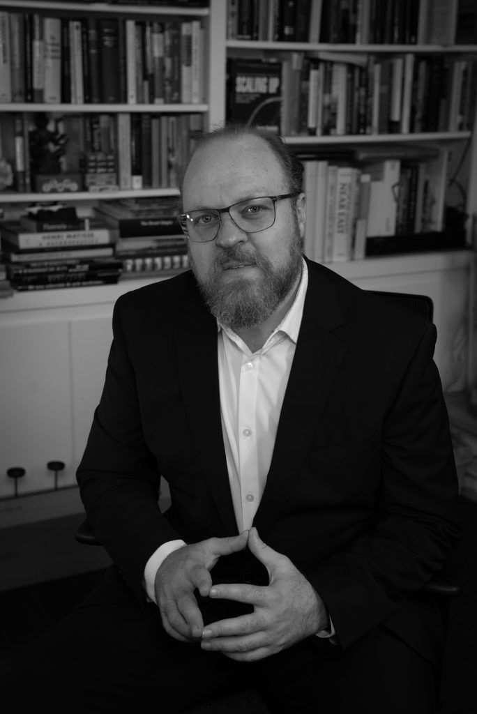 Black and white photo of Max Kozlovsky seated in front of a bookshelf in a dark jacket and open-collar white shirt, hands together, listening.